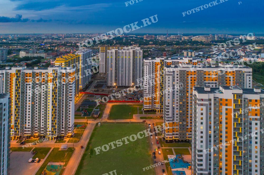 На фотографии изображены новые жилые кварталы в Санкт-Петербурге, снятые с высоты птичьего полета. Высотные дома с яркими фасадами освещены вечерним светом, создавая контраст с окружающим городским ландшафтом. 
Это изображение можно использовать в материалах по недвижимости, городскому планированию, архитектуре, а также в рекламных и информационных публикациях о жизни в современном мегаполисе.